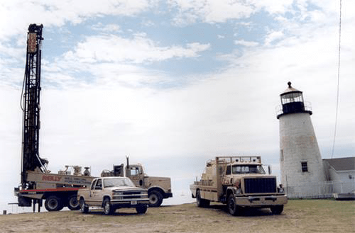 (from background to foreground) A drilling truck, a pickup truck, and a semi-trailer truck parked in front of a lighthouse